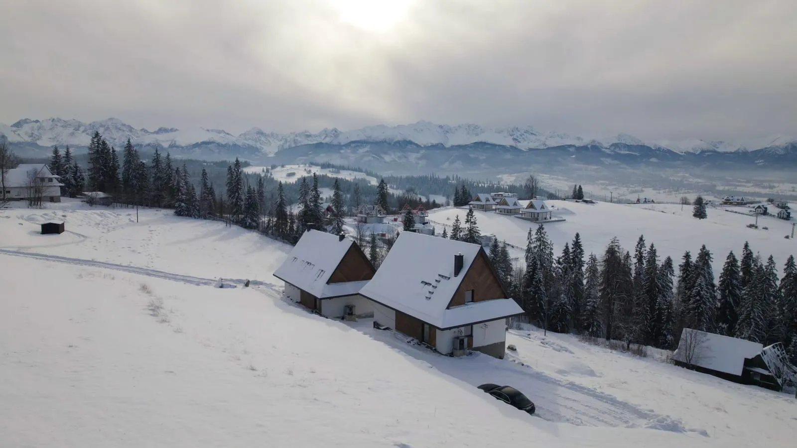 Zimowa panorama z drona, dwa domy w Gliczarowie Górnym na zaśnieżonym stoku, za nimi świerkowy las i Tatry