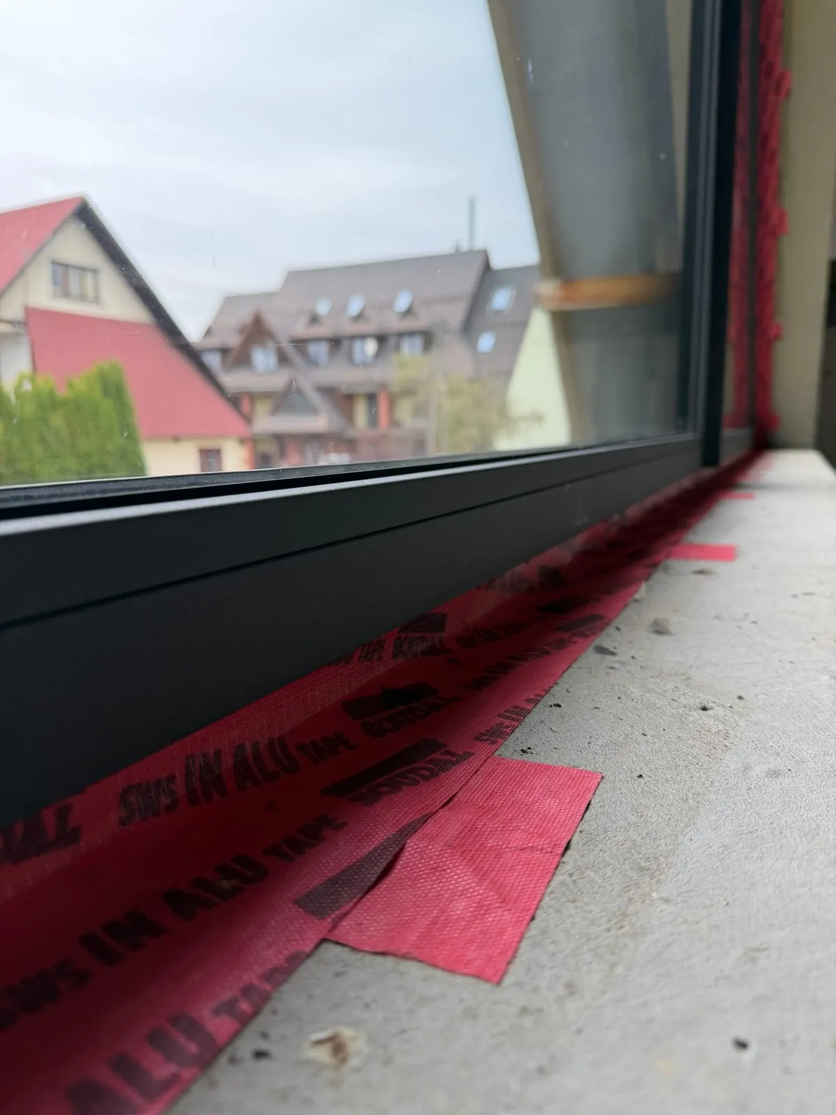 Dół okna od środka, czerwona taśma paroszczelna Soudal SWS na podokienniku, za szybą sąsiednie domy