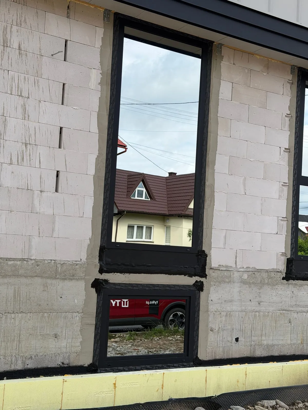 Wysokie okno z lustrzaną szybą, w odbiciu sąsiednie domy i bus TYLKA